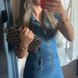 Pull&Bear Byxdress  - Säljer en blå jeansklänning med korta ärmar, markerad midja och hel dragkedja framtill. Klänningen har klassisk skjortkrage, fickor och figurnära passform. Perfekt för dig som gillar en cool och avslappnad denimlook.