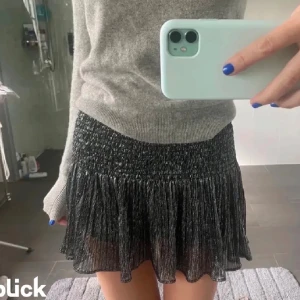 Glittrig svart kortkjol med volang - Supersnygg svart kortkjol med glittrigt tyg och volang nertill. Kjolen har smockad midja som sitter bekvämt och ger en cool look. Perfekt för dig som vill sticka ut lite extra med en trendig och festlig vibe.