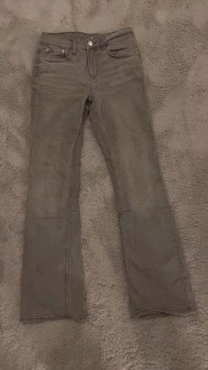 Grå bootcut jeans - Snygga grå jeans med bootcut passform och klassisk femficksdesign. Jeansen har en knapp och dragkedja framtill samt subtila slitningar för en cool look. Perfekta för dig som gillar en lite bredare benslut och vill ha en stilren vibe.