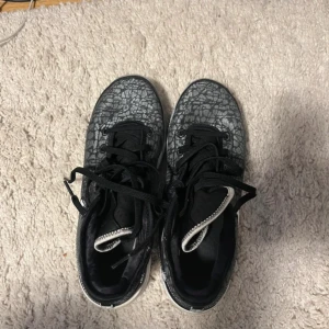 Nike sneakers med grått mönster kartong finns - Säljer ett par snygga Nike sneakers med grått och svart grafiskt mönster över hela ovandelen. Skorna har vit sula med detaljerad struktur undertill och en stor vit Swoosh på sidan med svart söm. Svarta skosnören och låg profil gör dem riktigt stilrena.