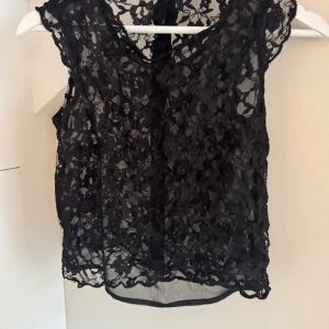 Svart blommig spetsblus med knappar - Säljer en svart blus i transparent spets med blommigt mönster. Blusen är ärmlös och har dekorativa knappar längs ryggen. Perfekt för dig som gillar detaljer och vill ha en cool, feminin vibe. Passar till både jeans och kjol.