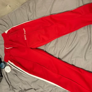 Palm Angels röda mjukisbyxor med vita ränder - Palm Angels röda mjukisbyxor med vita sidoremsor och broderad logga på fickan. Byxorna har elastisk midja med snörning och är tillverkade i polyester för en skön och sportig känsla. Perfekt för en chill och trendig streetwear-look.