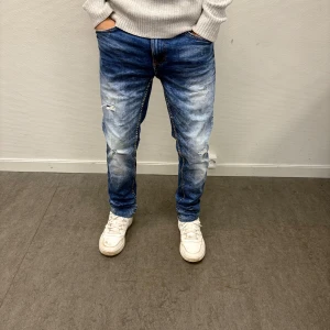 Blå Jeans från SMOG - Snygga blå jeans med slitningar och ljusa tvättade partier. Modellen är skinny fit med normal midja och klassiska fem fickor. Jeansen har en modern look med lite trasiga detaljer på benen och är tillverkade i stretchigt denimtyg för skön passform. Perfekt för dig som vill ha en clean och stilren look!