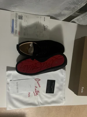 Christian Louboutin svarta sneakers - Svarta sneakers från Christian Louboutin med klassisk röd sula och broderad logga på hälen. Skorna har rund tå, snörning och är tillverkade i mocka med skinnfoder. Snygg och stilren design med exklusiv känsla, levereras med dustbag och äkthetskort. Passar både 41-42.