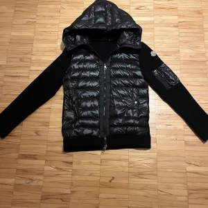 Svart Moncler pufferjacka med huva - Säljer en svart pufferjacka från Moncler med stickade ärmar och vadderad kropp. Jackan har en stor huva, dragkedja framtill och Moncler-logga på ärmen. Snygg och sportig look med fickor och detaljer i blankt material.