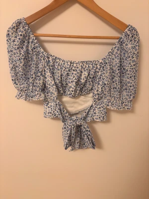Blommig croppad blus med knyt - Supersöt blommig blus i vitt och blått med puffiga korta ärmar och stor rosett i ryggen. Toppen är croppad och har öppen rygg med knytband som ger en snygg siluett. Den är från New Yorker (Berlin) och bara använd ett fåtal gånger. Modellen bär storlek S. Tveka inte att fråga. 💞