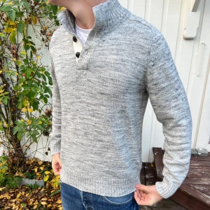 Grå stickad tröja med knappar - Snygg grå stickad tröja med ribbad hög krage och tre svarta knappar framtill. Tröjan har långärmad passform och är tillverkad i mjukt stickat material som ger en chill och stilren look. Perfekt att bära över en t-shirt för extra lager. Den har en lagning i ena armhålan men är inget märkvärdigt. Annars helt felfri och redo för en ny köpare! Skriv vid funderingar🙌🤝