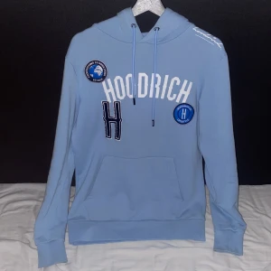Ljusblå hoodie från Hoodrich - Snygg ljusblå hoodie från Hoodrich med vita och blå broderade patches och tryck på bröstet. Tröjan har huva med snörning, känguruficka och ribbade muddar. Perfekt för dig som gillar streetwear och vill sticka ut med coola detaljer.