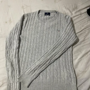 Grå kabelstickad tröja från GANT - Snygg ljusgrå kabelstickad tröja från GANT i stretchig bomull. Rund halsringning och diskret broderad logga på bröstet. Tröjan har långa ärmar och ribbade muddar vid ärmslut och nederkant. Perfekt för dig som gillar klassisk och clean stil.