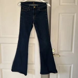 Mörkblå bootcut jeans från Lee - Snygga mörkblå jeans från Lee med klassisk femficksdesign och kontrastsömmar. Modellen har bootcut-ben och normal midja, vilket ger en retrovibe. Jeansen är tillverkade i mjukt denimtyg och har Lee-logga på bakfickan och läderpatch i midjan. Strectch. 