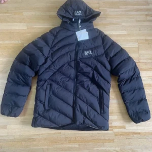 Svart dunjacka från EA7 Emporio Armani - Säljer en svart dunjacka från EA7 Emporio Armani med huva och snygga diagonala quiltade sömmar. Jackan har dragkedja framtill, två sidofickor och EA7-logga på bröstet och huvan. Perfekt för kalla dagar och har en modern, sportig look.