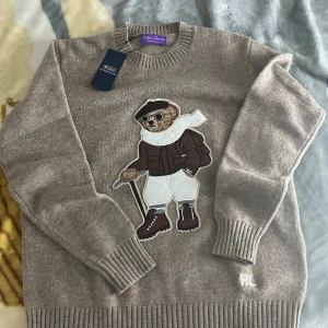 Beige stickad tröja Polo Bear Ralph Lauren - Beige stickad tröja från Polo Ralph Lauren med ikonisk Polo Bear-applikation framtill. Tröjan har ribbade muddar vid hals, ärmslut och nederkant samt långärmad passform. Tillverkad i mjuk ull och har en klassisk rund halsringning.