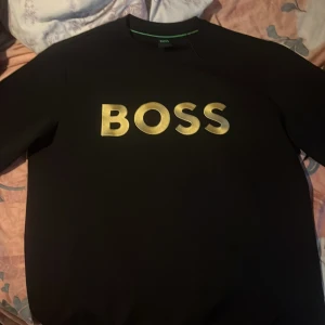 Svart BOSS sweatshirt med guldtryck - Svart sweatshirt från BOSS med stort, glansigt guldtryck över bröstet. Tröjan har en klassisk rund hals och långa ärmar. Perfekt för dig som gillar stilrena och trendiga plagg med tydlig logga. Den är helt ny och aldrig använd säljer den för den var stor när jag köpte den