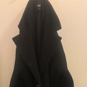 Svart kappa från ONLY med bälte - Säljer en svart kappa från ONLY med bred krage och knappar framtill. Jackan har ett matchande bälte i midjan och långa ärmar. Perfekt för dig som gillar stilrena och klassiska plagg med snygg siluett.