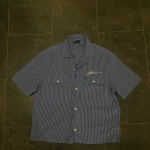 Blåvit randig skjorta från Soulland - Snygg kortärmad skjorta från Soulland i blått och vitt med tunna ränder. Skjortan har två bröstfickor med knappar, klassisk krage och vit broderad logga på vänster sida. Tillverkad i mjuk bomull och har en loose fit som ger chill vibe.