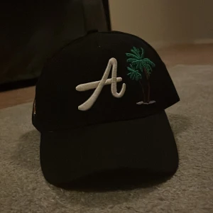 Svart keps med palmträd och LA-brodyr - Svart keps med böjd skärm, broderat stort vitt A och två gröna palmträd framtill. På sidan finns en broderad glob med texten 'Los Angeles AR 1989' i guld och rött. Snygg streetstil och perfekt för sommaren.