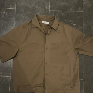 Brun kortärmad skjorta från SÉFR - Snygg brun skjorta från SÉFR med korta ärmar och två stora bröstfickor. Skjortan har klassisk krage och knäppning framtill med matchande knappar. Tillverkad i bomull, perfekt för dig som gillar minimalistisk och clean stil.