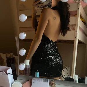 Svart glittrig paljettklänning - Supersnygg svart kort klänning täckt av glittriga paljetter. Hög hals och öppen rygg ger en riktigt cool vibe. Tight passform som framhäver figuren och ärmlös design. Perfekt för dig som vill sticka ut och glänsa.