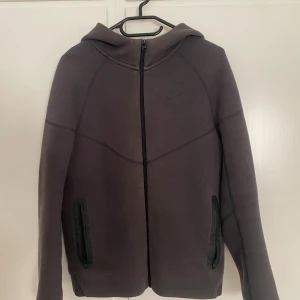Mörkgrå hoodie från Nike XL - Snygg mörkgrå hoodie från Nike i storlek XL, med hel dragkedja framtill och två svarta fickor med dragkedja. Tröjan har huva och långa ärmar, perfekt för chill eller träning. Diskret Nike-logga på bröstet och stilrena sömmar ger en clean look.