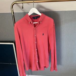 Rosa skjorta från Ralph Lauren M - Riktigt snygg rosa skjorta från Ralph Lauren i storlek M. Skjortan har klassisk krage, knappar hela vägen och lång ärm. Materialet är lätt mesh-bomull som känns skönt mot huden. På bröstet finns den ikoniska blå Ralph Lauren-loggan broderad.