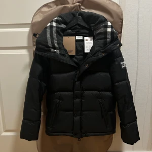 Burberry Jacka/Väst - Svart pufferjacka från Burberry med klassisk rutig huva i grå toner. Jackan har dragkedja och knappar framtill, två fickor med dragkedja och en patch med Burberry London England på ärmen. Perfekt för kalla dagar med sin dunfyllda design och snygga detaljer.