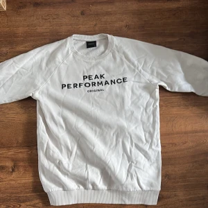 Vit Peak Performance sweatshirt - Vit sweatshirt från Peak Performance med svart broderad logga på bröstet. Klassisk rund halsringning och långa ärmar. Mjuk insida och ribbade muddar vid ärmslut och nederkant. Perfekt för en chill och sportig stil.