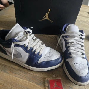 Nike Air Jordan 1 Low vit/marinblå - Säljer ett par Nike Air Jordan 1 Low i vit och marinblå färg. Skorna har klassisk snörning, rund tå och är tillverkade i skinn med textildetaljer. Ikonisk Air Jordan-logga broderad på hälen och Jumpman på plösen. Perfekta för dig som gillar streetwear och sneakers med retrovibe.