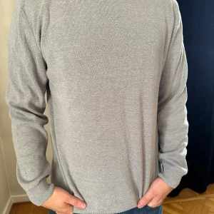 Ljusgrå stickad tröja med rund hals - En stilren ljusgrå stickad tröja med rund hals och ribbade muddar. Tröjan har en klassisk passform och är perfekt för lager-på-lager. Enkel design utan mönster eller logga, passar till jeans eller chinos.