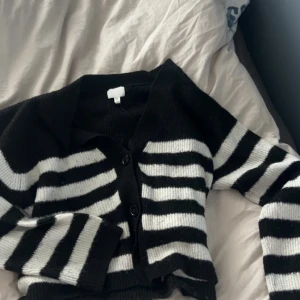 Randig svartvit stickad kofta - Säljer en stickad kofta med svarta och vita breda ränder. Koftan har knappar framtill, krage och långa ärmar. Den är ribbstickad och har en klassisk, lite boxig passform. Perfekt för dig som gillar en stilren men ändå edgy look.
