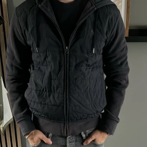 Svart pufferjacka från Polo Ralph Lauren - Svart pufferjacka från Polo Ralph Lauren med huva och dragkedja framtill.