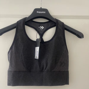 Mörkgrå sporttopp från 157 - Sportig mörkgrå topp från 157 med ribbade partier i nederkant och sidor. Modellen är ärmlös med brottarrygg och har en tight passform som sitter skönt vid träning. Materialet är stretchigt och mjukt, perfekt för aktiva dagar.