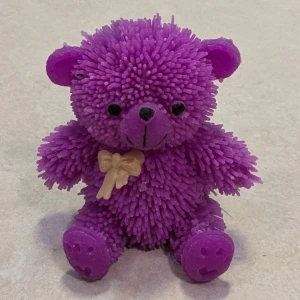Jätte söt lila björn  - Lila gummibjörn med piggig textur och en beige rosett. Den är mjuk, stretchig och passar som stressleksak eller dekoration. . Produkten visar inget tydligt slitage eller defekter.