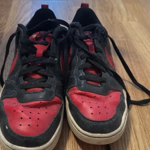 Nike sneakers svart/röd med snörning - Snygga Nike sneakers i svart och rött med klassisk swoosh på sidan. Skorna har lågt skaft, svart snörning och perforerad tå. Ovandelen är i skinn och syntet, med vit sula och röda detaljer på häl och plös. Perfekta för dig som gillar streetstyle.