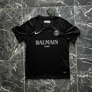 Svart PSG x Balmain fotbollströja Nike - Snygg svart fotbollströja från Nike i samarbete med PSG och Balmain. Tröjan har korta ärmar, rund hals och är tillverkad i Dri-FIT-material som andas. Framsidan pryds av Balmain Paris-logga och PSG-emblem. Perfekt för dig som vill sticka ut på planen.