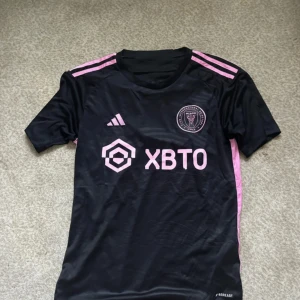 Inter Miami CF svart fotbollströja XS - Svart Inter Miami CF matchtröja från Adidas med rosa detaljer och logga på bröstet. Tröjan har korta ärmar, rund hals och är tillverkad i ett lätt, tekniskt material som andas. Rosa Adidas-ränder på axlarna och sponsortryck framtill.