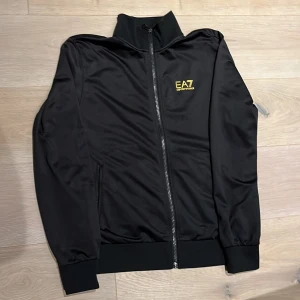 Svart polyester kofta från EA7 Emporio Armani - Svart kofta från EA7 Emporio Armani med gul logga på bröstet och detaljer i gult på ryggen. Jackan har hel dragkedja framtill, hög krage och ribbade muddar. Tillverkad i mjuk polyester för en sportig och clean look.