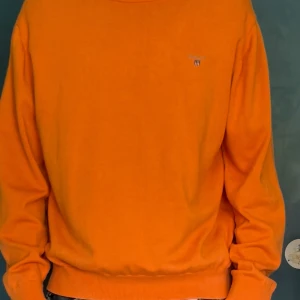Orange tröja från Gant - Snygg orange tröja från Gant med liten broderad logga på bröstet. Tröjan har rund halsringning, långa ärmar och ribbade muddar vid ärmslut och nederkant. Perfekt för dig som vill sticka ut med färg.