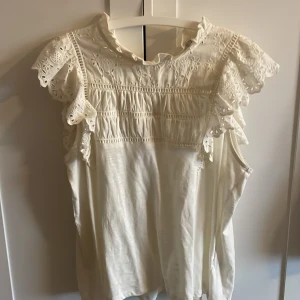 Vit blus med broderade volangärmar - Supersöt vit blus från Holly & Whyte med broderade detaljer och volangärmar. Blusen har rundad hals med liten volangkant och knäppning bak. Använd Max 1-2 gånger 