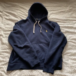 Marinblå hoodie från Polo Ralph Lauren - Säljer en marinblå hoodie från Polo Ralph Lauren med klassisk gul broderad logga på bröstet. Tröjan har huva med snörning, känguruficka och ribbade muddar. Perfekt för dig som gillar stilren och bekväm streetwear. Pris kan diskuteras!!