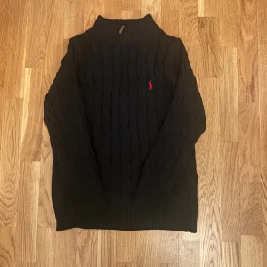 Svart kabelstickad tröja från Ralph Lauren - Svart kabelstickad tröja från Ralph Lauren med klassisk röd broderad logga på bröstet. Tröjan har ribbade muddar, högre krage och är långärmad. Perfekt för kyliga dagar och enkel att matcha med jeans eller kjol.