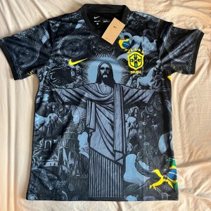 Nike Brasil jesus fotbollströja - Unik svart Brasilien fotbollströja från Nike med grafiskt tryck av Kristusstatyn och brasilianska symboler. Tröjan har korta ärmar, broderat CBF-märke och gula detaljer. Tillverkad i Dri-FIT-material som andas och passar perfekt för fotboll eller streetwear. Storlek L men kan även passa M