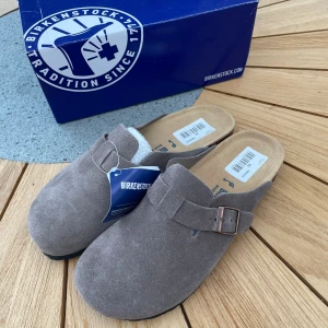 Grå mocka sandaler - Birkenstock sandaler i grå mocka med mjuk fotbädd och justerbart spänne i metall. Klassisk slip-on modell med rund tå och platt sula, perfekt för chill dagar. Snygg och enkel design som funkar till det mesta.