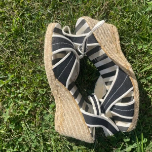 Randiga kilklackade sandaler - Säljer ett par sandaler med svartvita ränder och snygga snörningar runt ankeln. Skorna har hög kilklack i rep och öppen tå. Tillverkade i bomullstyg med vita detaljer. Perfekta för sommaren och ger en cool, avslappnad vibe.