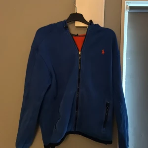 Blå fleecejacka från Polo Ralph Lauren - Blå fleecejacka med huva från Polo Ralph Lauren, med orange broderad logga på bröstet och orange insida. Jackan har dragkedja framtill och praktiska fickor med zip. Perfekt för kyliga dagar och chill stil. PRISET KAN DISKUTERAS!! 