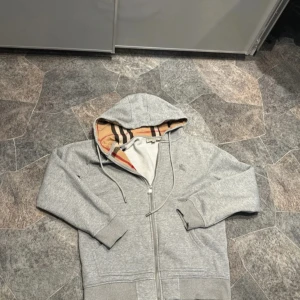Grå hoodie från Burberry med rutig huva - Snygg grå hoodie från Burberry med klassiskt rutigt mönster i beige, svart och rött på insidan av huvan och öppningen. Tröjan har dragkedja, snörning vid huvan och broderad Burberry-logga på bröstet. Perfekt för dig som gillar exklusiva streetwear-vibbar.