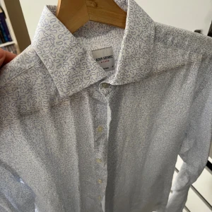 Vit skjorta med blått mönster John Henric - Snygg vit skjorta från John Henric med ljusblå mönster. Size 38, vilket motsvarar Small. Tillverkad i italienskt bomullstyg som känns mjukt och fräscht mot huden. Perfekt för dig som gillar stilrena detaljer.