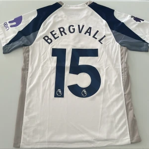 Tottenham Bergvall #15 Nike fotbollströja - Säljer en Tottenham Hotspur fotbollströja med Bergvall och nummer 15 på ryggen. Tröjan är vit med marinblå och grå detaljer, röd AIA-logga på bröstet och Premier League-märke på ärmen. Tillverkad i lätt och ventilerande material, perfekt för match eller träning. Replika, endast handtvätt gäller!