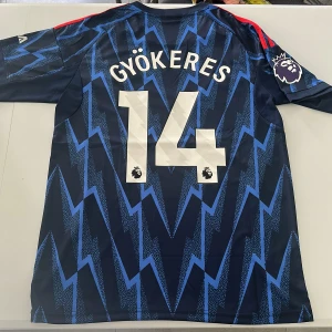 Arsenal bortatröja Gyökeres #14 Adidas - Snygg Arsenal bortatröja från Adidas med Gyökeres och nummer 14 på ryggen. Tröjan är marinblå med blått blixtmönster, röda axelränder och Premier League-logga på ärmen. Tillverkad i lätt och ventilerande polyester, perfekt för match eller träning. Replika, endast handtvätt gäller!