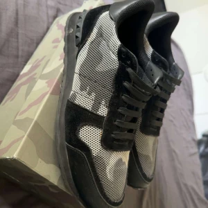 Valentino Garavani camo sneakers - Säljer ett par Valentino Garavani sneakers med svart och grått camouflagemönster. Skorna har svarta mockadetaljer, svarta snören och klassiska nitar på hälen. Ovandelen är i mesh och skinn för en lyxig känsla. Kommer med originalkartong och röd dustbag. Skorna är storlek 44 men passar 43.