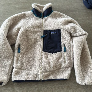 Beige teddyjacka från Patagonia XXS - Mysig teddyjacka från Patagonia i beige med marinblå detaljer och dragkedjor. Jackan har hög krage, tre fickor med dragkedja och en kontrastficka på bröstet. Perfekt för kyliga dagar och riktigt skön att ha på sig.